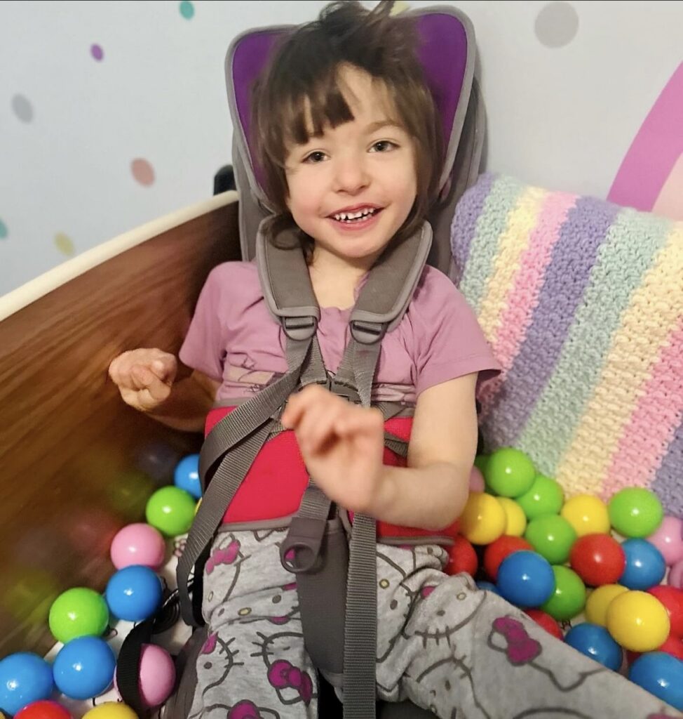 A young girl seated in a SleepSafe bed with a lot of colorful balls surrounding her.