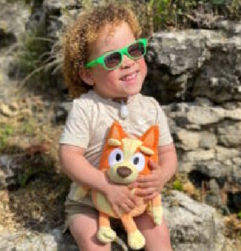A young child wearing sunglasses holding a stuffed animal and sitting on some rocks.