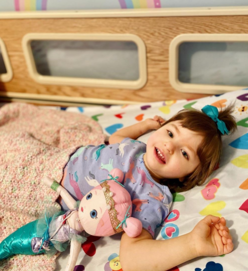 A young girl lying in a SleepSafe bed with a doll beside her.